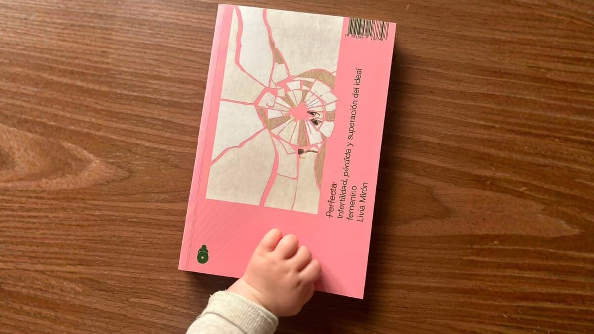 Una mano de bebé se acerca al libro Perfecta sobre una mesa de madera, en una imagen del artículo de Infobae